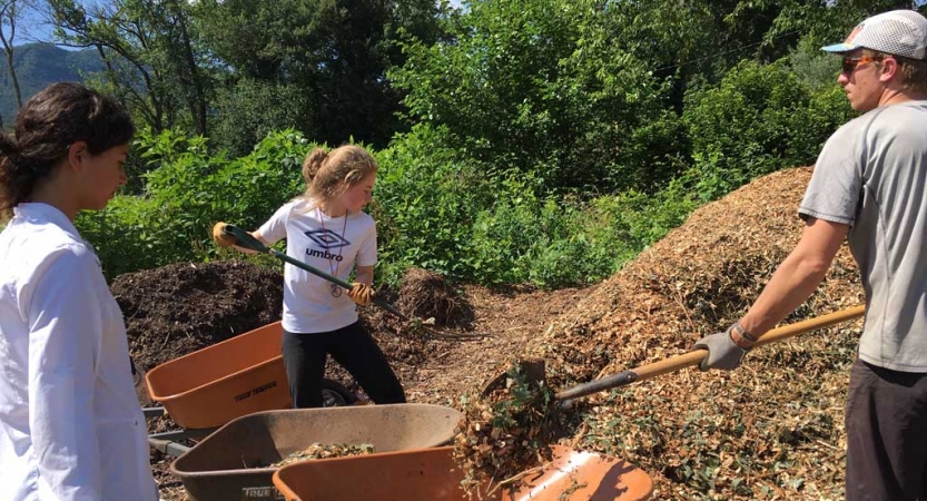 Two people shovel mulch into a wheelbarrow, held by another. 